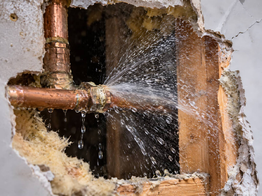 Burst copper pipe spraying water inside a wall.