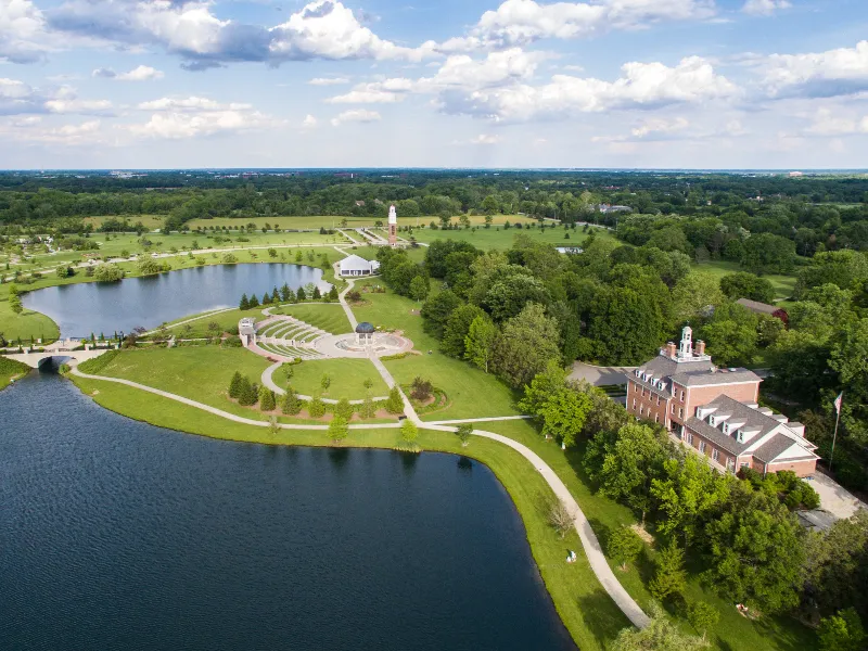 Aerial view of a park with a lake, walking paths, amphitheater, and a large brick building surrounded by trees.