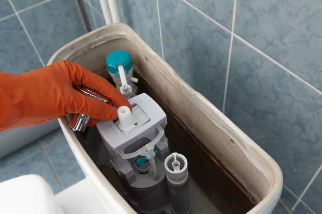 Hand in orange glove repairing or adjusting the inside mechanism of a toilet tank.