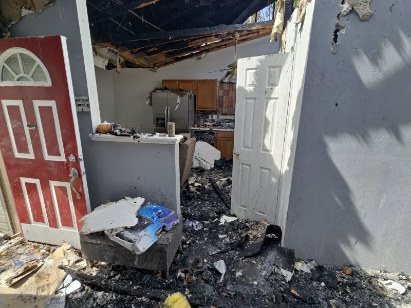 Interior of a house with fire damage, showing a burnt floor, damaged walls, and a partially collapsed ceiling.