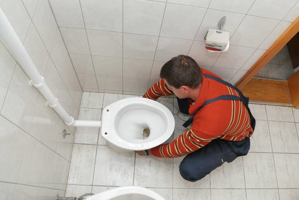 A man in a striped sweater installs or repairs a toilet bowl in a tiled bathroom.