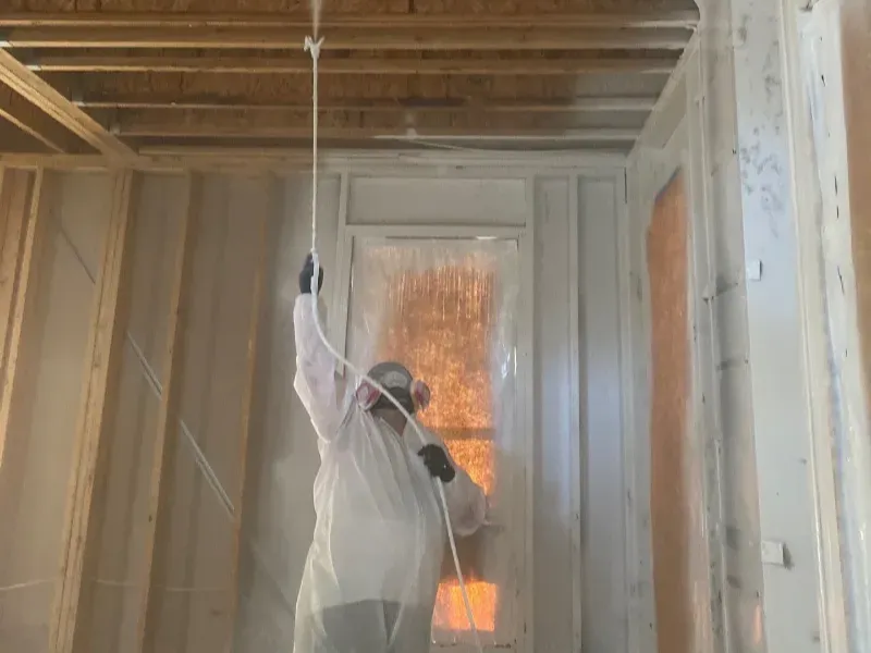 Person in protective suit and mask spraying insulation foam on walls and ceiling inside a building.