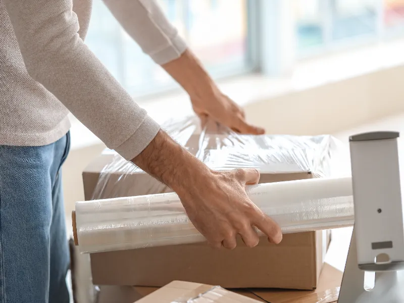 Person wrapping a cardboard box with clear plastic wrap on a table.