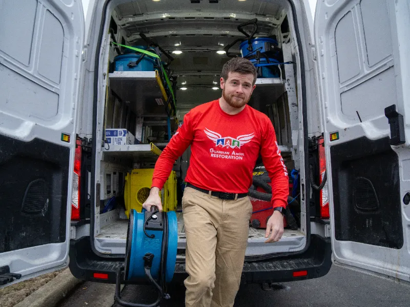 Man in red shirt carrying blue equipment out of a white van with open back doors.